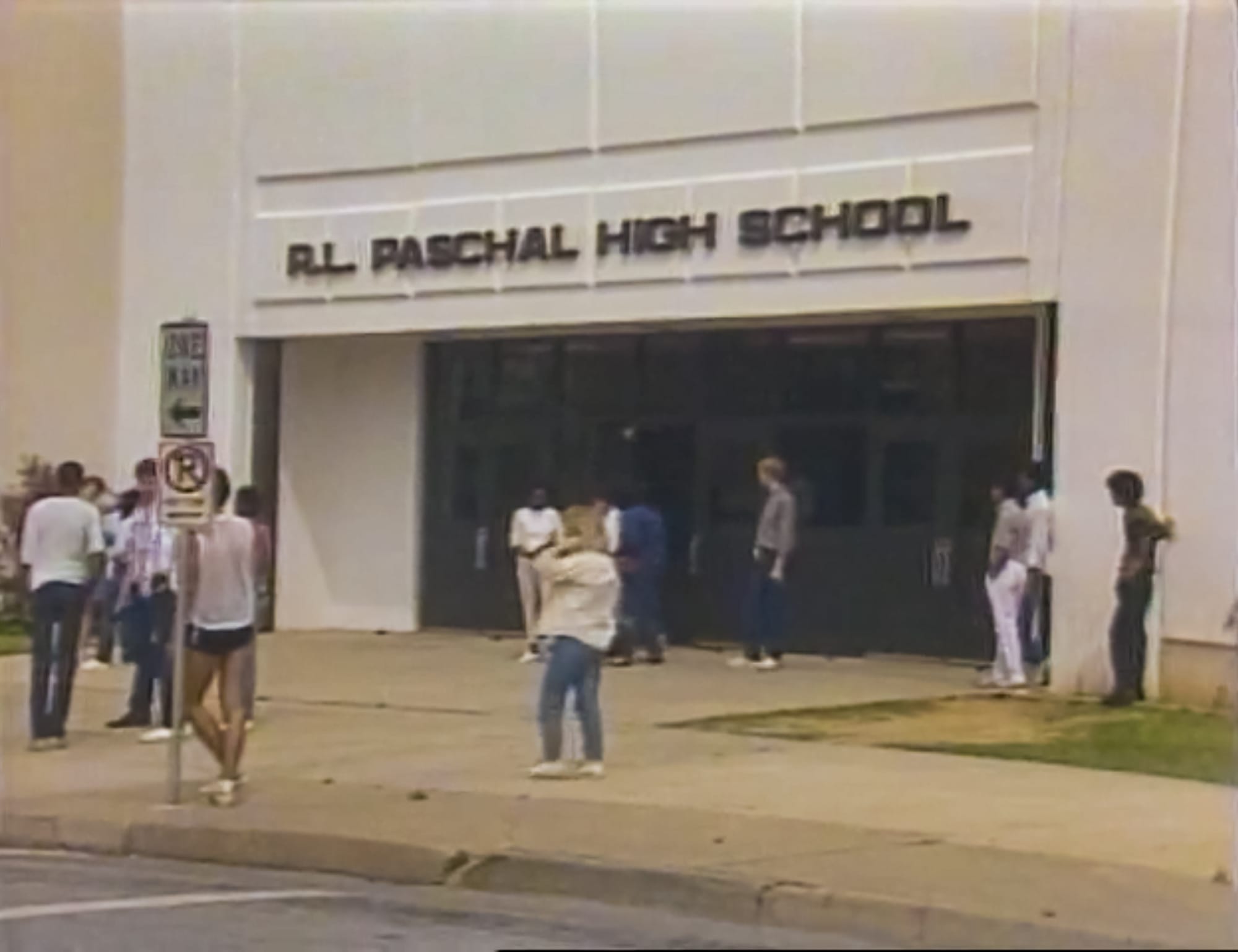 Students stand around a plain beige wall with inset glass doors and windows.